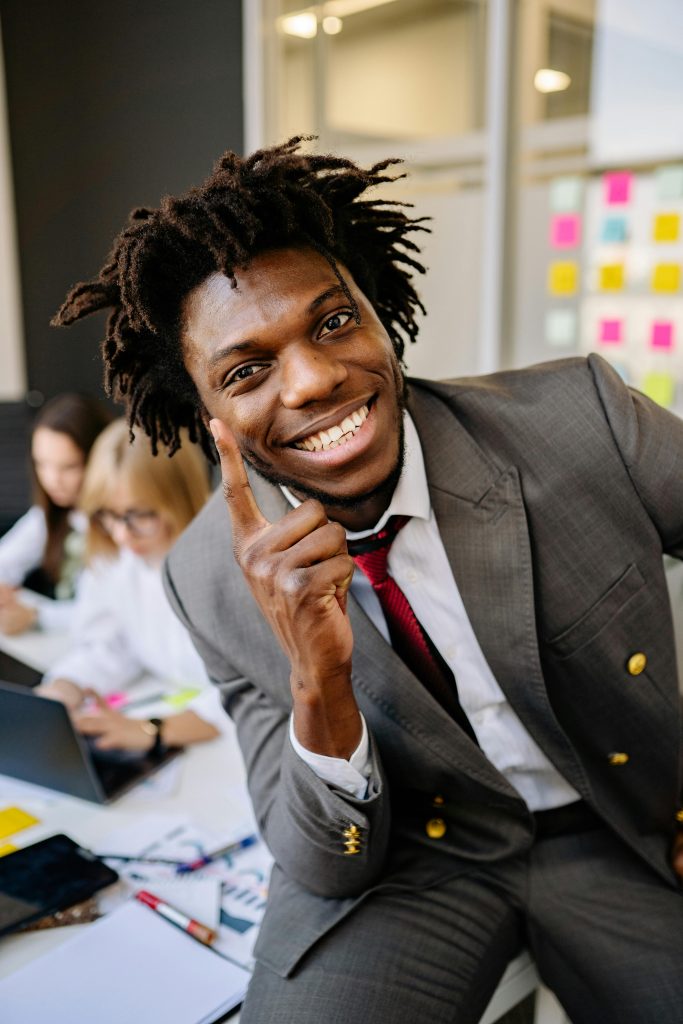 Smiling businessman in a gray suit points finger, exuding confidence and positivity in an office setting.